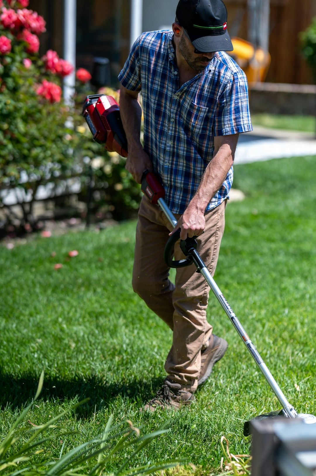Man in plaid shirt trims grass in sunny backyard.
